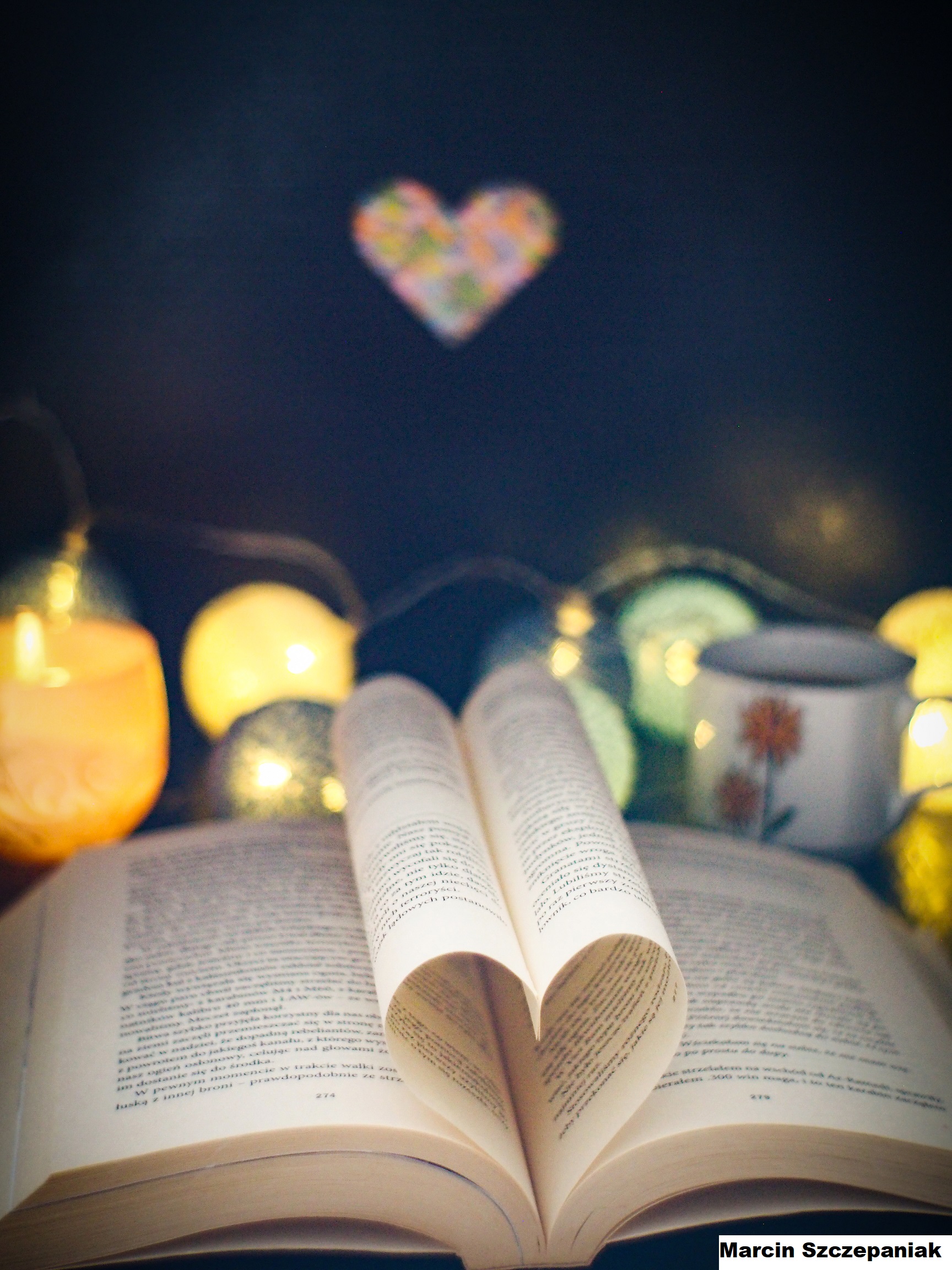 Zdjęcie przedstawia rozłożona książkę, w której środkowe kartki ułożone są w kształcie serca. W tle widać palące się lampki i światełka. 