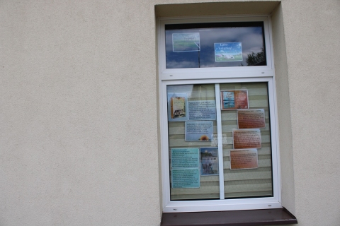 Zdjęcie prezentuje wystawę pt. „Książki z latem w tle” na oknach Powiatowej Biblioteki Publicznej, która prezentuje książki, których akcja rozgrywa się latem, a wakacyjne miesiące stanowią tło opowiadanych historii.