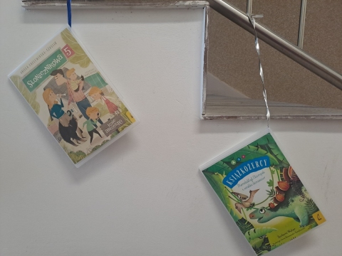 Zdjęcie prezentuje wystawę powieszoną na korytarzu przed biblioteką prezentującą różne okładki książek dla dzieci w których głównymi bohaterami są literackie dinozaury.