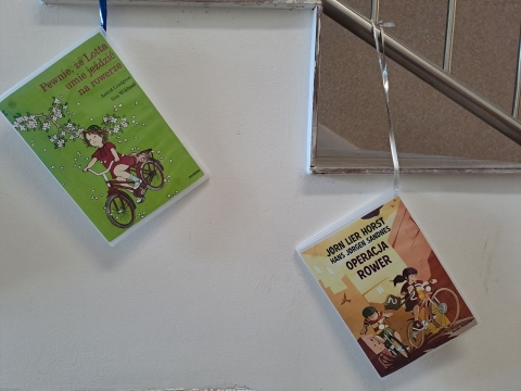 Zdjęcie prezentuje wystawę powieszoną na korytarzu przed biblioteką prezentującą różne okładki książek dla dzieci, w których ważnym punktem jest jazda na rowerze.  