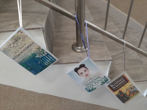 Zdjęcie prezentuje wystawę powieszoną na korytarzu przed biblioteką prezentującą różne okładki książek z wiosną w tytule.