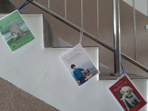 Zdjęcie prezentuje wystawę powieszoną na korytarzu przed biblioteką prezentującą różne okładki książek w których głównym bohaterem jest pies.