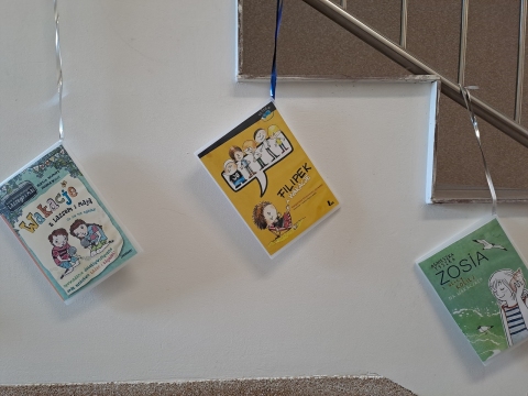 Zdjęcie prezentuje wystawę powieszoną na korytarzu przed biblioteką prezentującą różne okładki książek z wyrazem wakacje w tytule.