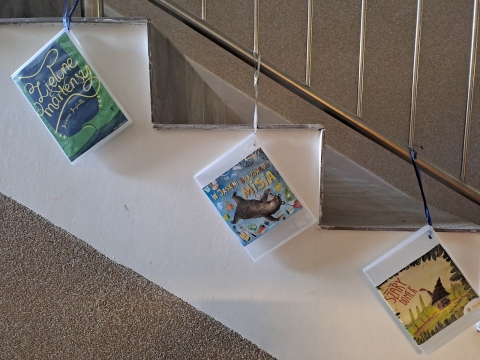 Zdjęcie prezentuje wystawę powieszoną na korytarzu przed biblioteką prezentującą różne okładki książek dla dzieci, w tytule których występuje nazwa jakiegoś koloru.  