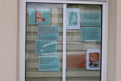Zdjęcie prezentuje wystawę pt. „Książki z latem w tle” na oknach Powiatowej Biblioteki Publicznej, która prezentuje książki, których akcja rozgrywa się latem, a wakacyjne miesiące stanowią tło opowiadanych historii.