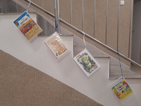 Zdjęcie prezentuje wystawę powieszoną na korytarzu przed biblioteką prezentującą różne okładki książek dla dzieci w których głównymi bohaterami są literackie dinozaury.