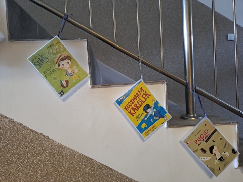 Zdjęcie prezentuje wystawę powieszoną na korytarzu przed biblioteką prezentującą różne okładki książek, w których bohaterami są dzieci. 