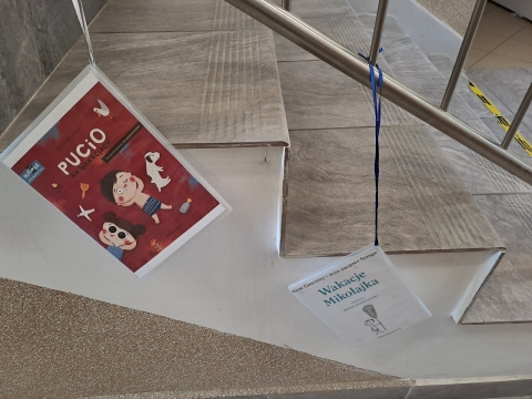 Zdjęcie prezentuje wystawę powieszoną na korytarzu przed biblioteką prezentującą różne okładki książek z wyrazem wakacje w tytule.