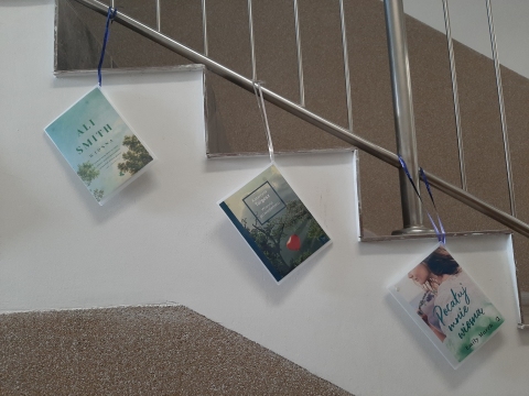 Zdjęcie prezentuje wystawę powieszoną na korytarzu przed biblioteką prezentującą różne okładki książek z wiosną w tytule.