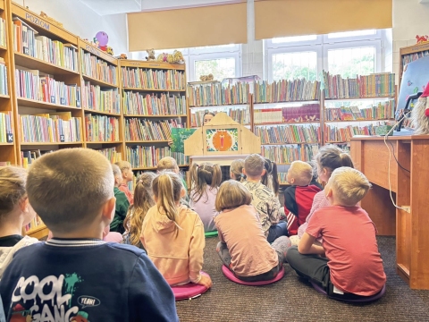 Zdjęcie prezentuje lekcję biblioteczną z cyklu "W magicznym świecie Kamishibai", która odbyła się 13 maja 2026 r.  dla grupy 5- i 6-latków z kazimierskiego przedszkola. Na zdjęcie widać siedzące w bibliotece dzieci, które patrzą w kierunku otwartego teatrzyku Kamishibai. 