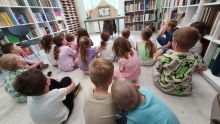 Zdjęcie prezentuje lekcję biblioteczną w klasie zerowej ze Szkoły Podstawowej nr 3 w Puławach. Na zdjęciu pani Katarzyna, dyrektor PBP w Puławach i uczniowie zerówki. Na zdjęciu dzieci siedzą tyłem i patrzą na otworzony teatrzyk Kamishibai.