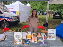 Zdjęcie przedstawia akcję bookcrossing, która odbyła się 12 lipca 2026 r. podczas Nadwiślańskich Spotkań Kulinarnych. Na zdjęciu stolik na którym ułożone są książki oraz dwa plakaty informujące o akcji i prezentujące logo biblioteki. W tem pani dyrekotr biblioteki Katarzyna Adamowska-Kane. 